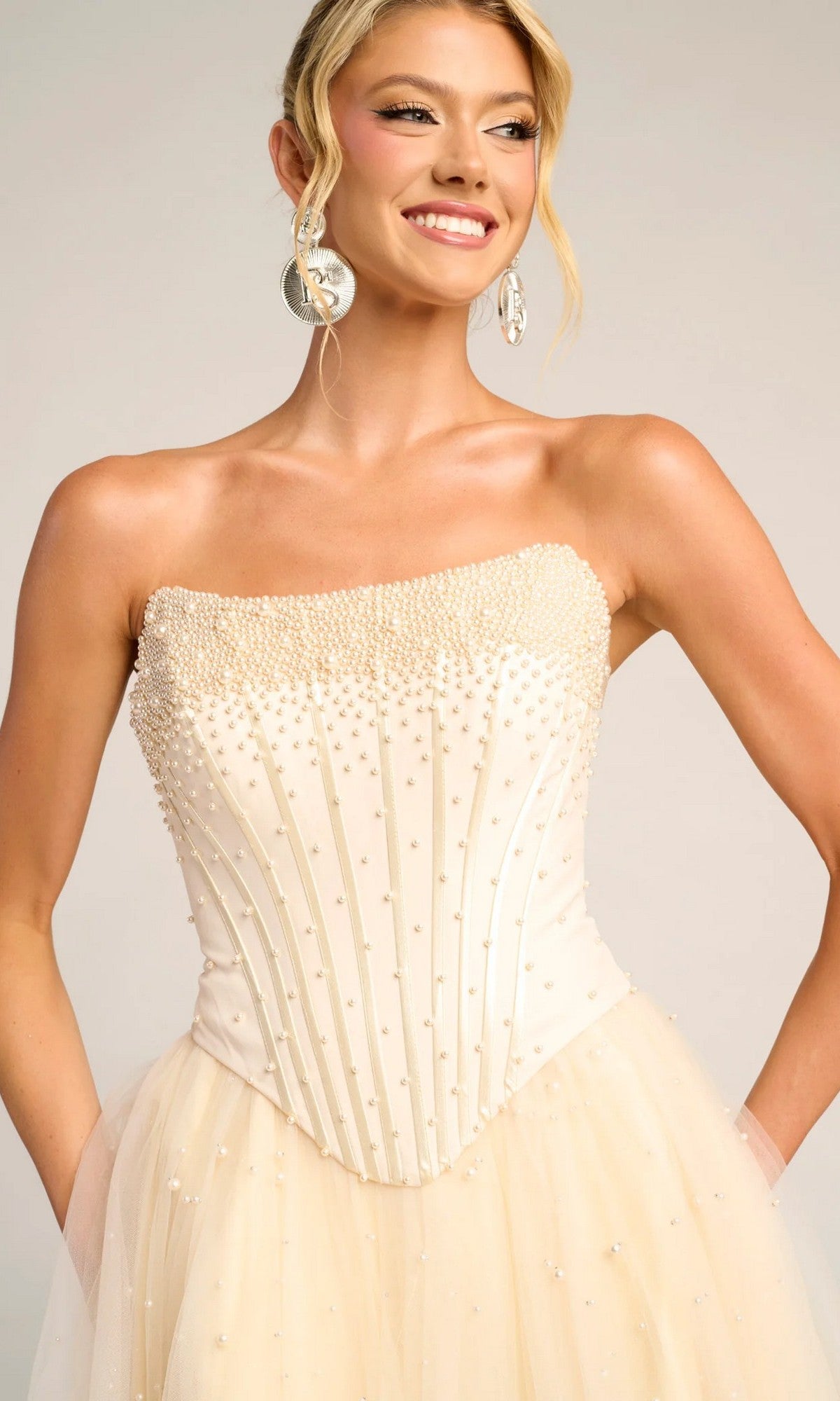 Woman wearing the Portia and Scarlett PS26034 long formal dress, featuring a strapless corset bodice with pearls and a glitter tulle skirt, posing with hands on hips and smiling.